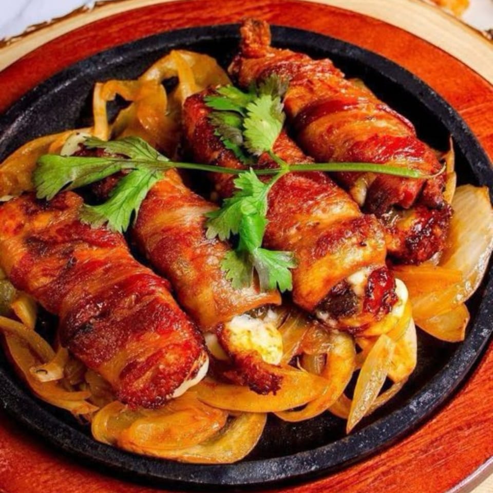 Bacon-wrapped shrimp and melted cheese on a sizzling skillet with sautéed onions, garnished with cilantro.