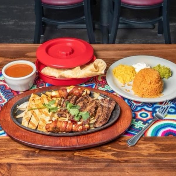 Sizzling fajitas with chicken and steak, rice, guacamole, sour cream, tortillas, and salsa on a colorful placemat.