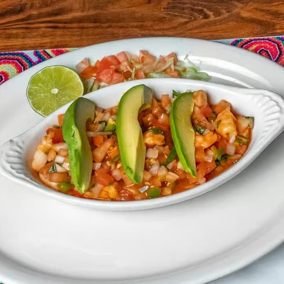 Shrimp ceviche with avocado slices, lime, and a side of lettuce and tomato on a white plate.