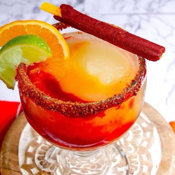 A vibrant red and orange Margarita with a chili-lime rim, garnished with orange, lime, and a tamarind candy.