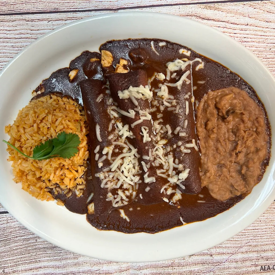 Three enchiladas covered in dark mole sauce and shredded cheese, with rice and refried beans.